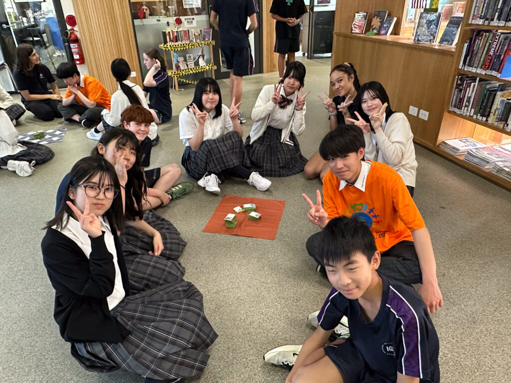 A group of pupils in school uniforms sit on the floor in a library, smiling and making peace signs around a board game—building connections through language and laughter. Bookshelves and more pupils are visible in the background.
