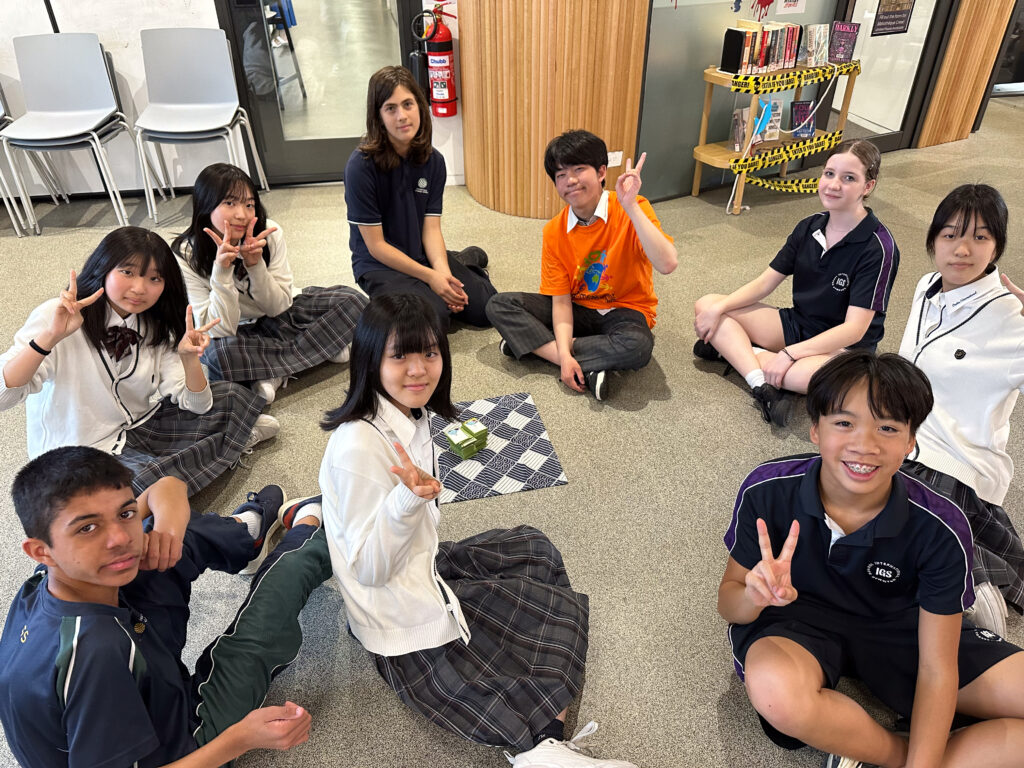A group of students sit in a circle on the floor, smiling, making peace signs, and building connections as they enjoy a board game or activity set. They are indoors near some chairs and wooden columns.