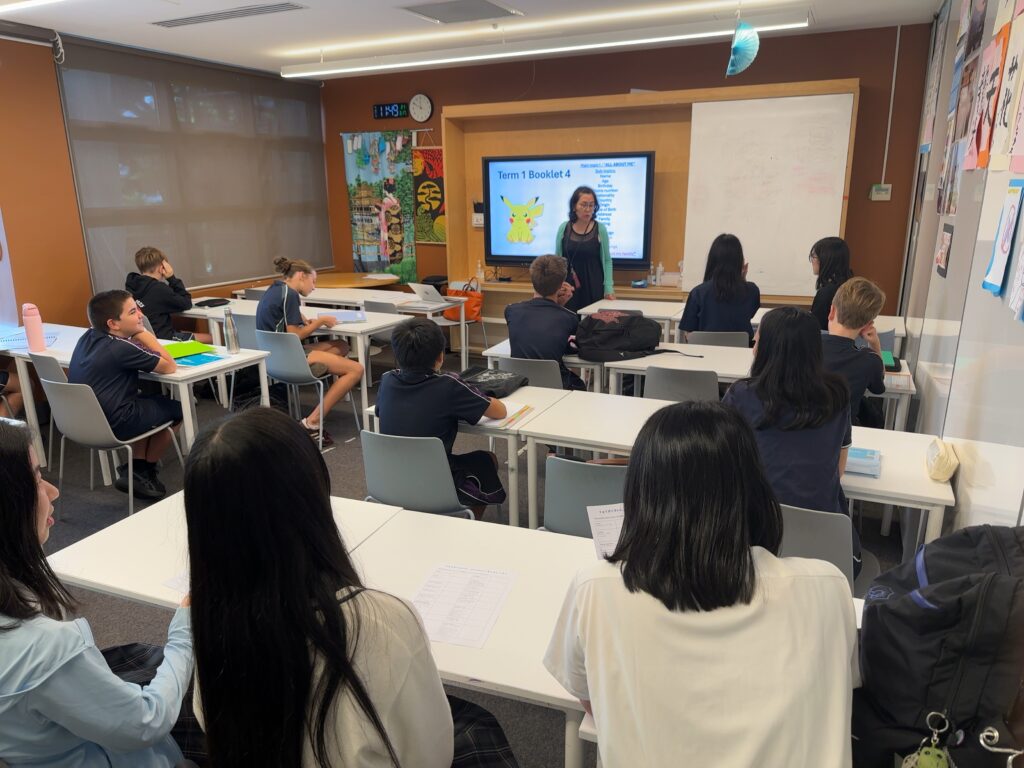 A teacher stands at the front of a classroom, using clear communication to engage attentive pupils. A whiteboard and large screen display information, whilst posters decorate the walls, fostering an environment for building connections.