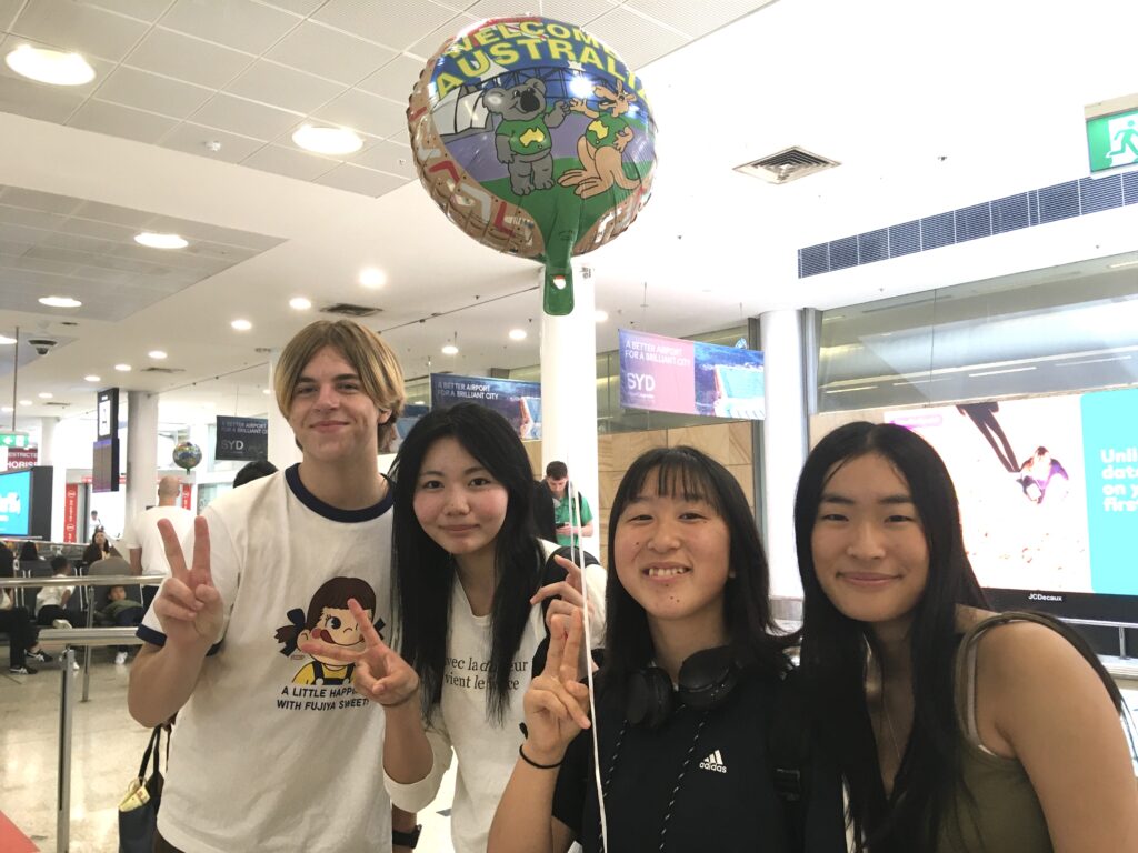 Four young people smile at the camera in an airport, one holding a colourful Welcome to Australia balloon. Two flash peace signs, building connections through shared language and excitement amid bright signs and bustling travellers.
