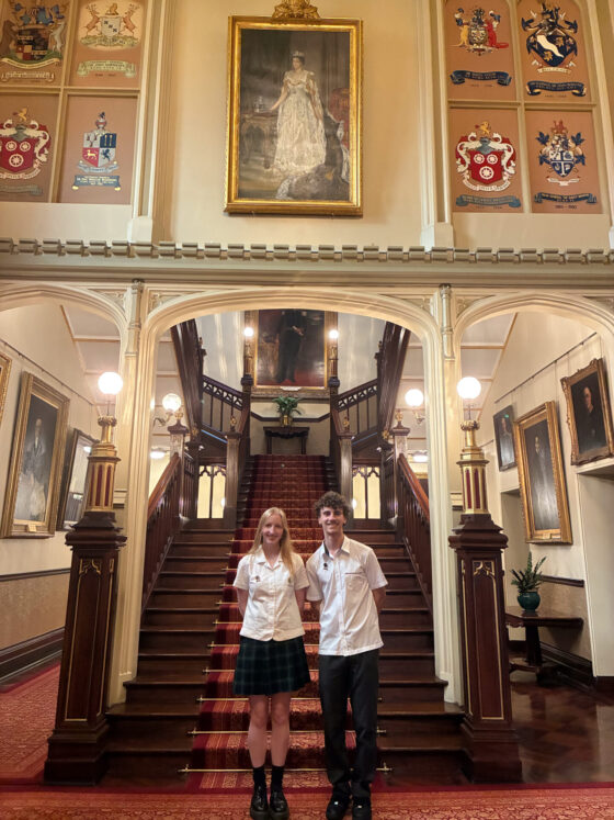 Two students in school uniforms stand at the foot of a grand staircase in an ornate hall, surrounded by portraits and coats of arms, with a large portrait of a woman in white above the stairs.