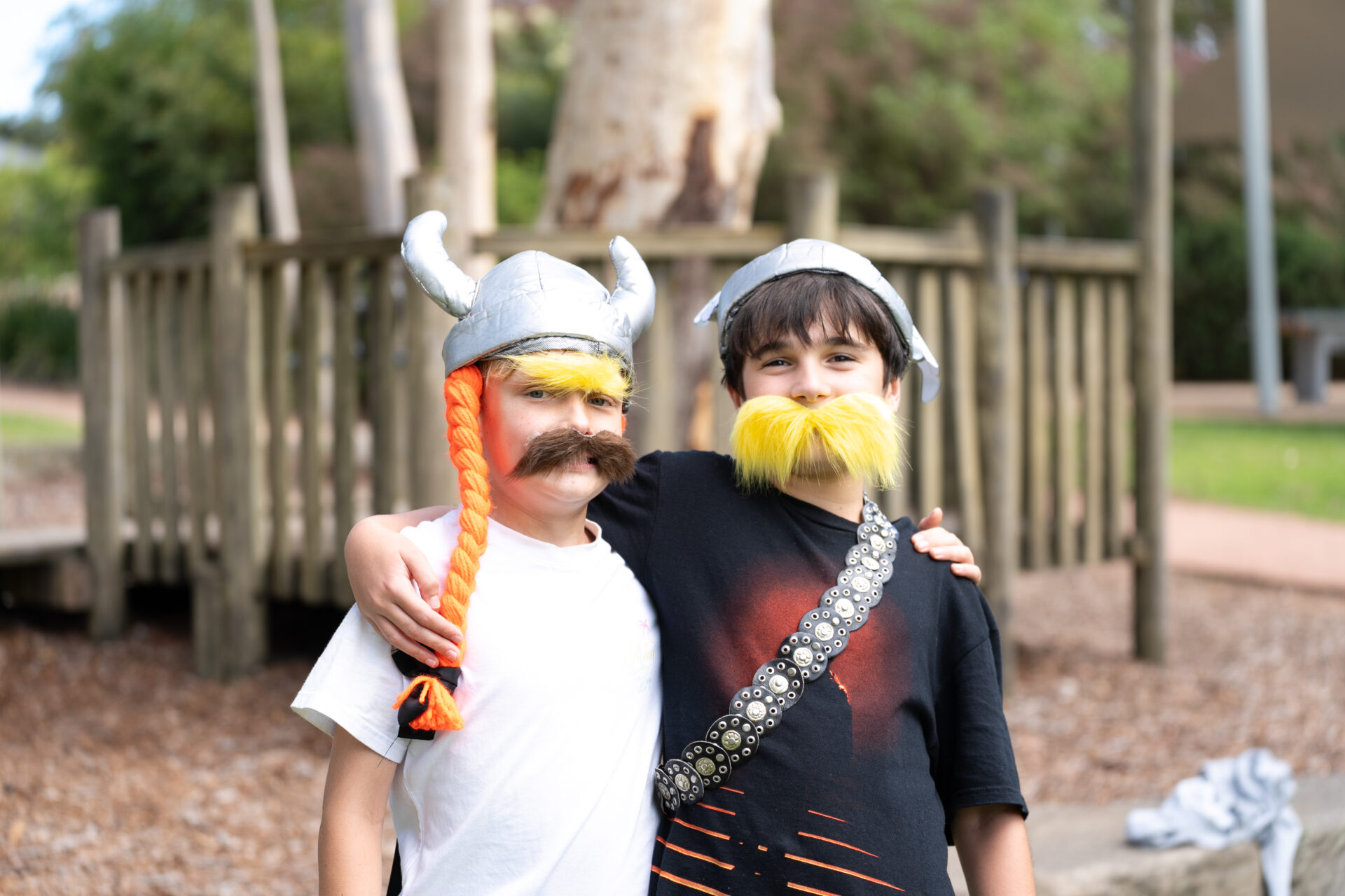 Two children dressed as Vikings, with horned helmets, fake moustaches, and yellow beards, stand arm-in-arm and smile outdoors in front of a wooden play structure.