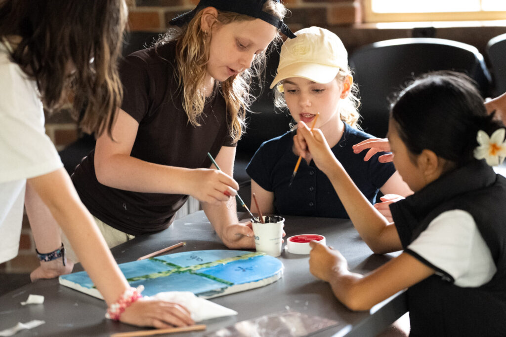 Four children sit around a table painting a craft project together, focused and engaged. Two girls hold paintbrushes whilst another dips her brush in a small pot. Sunlight streams in from a nearby window.