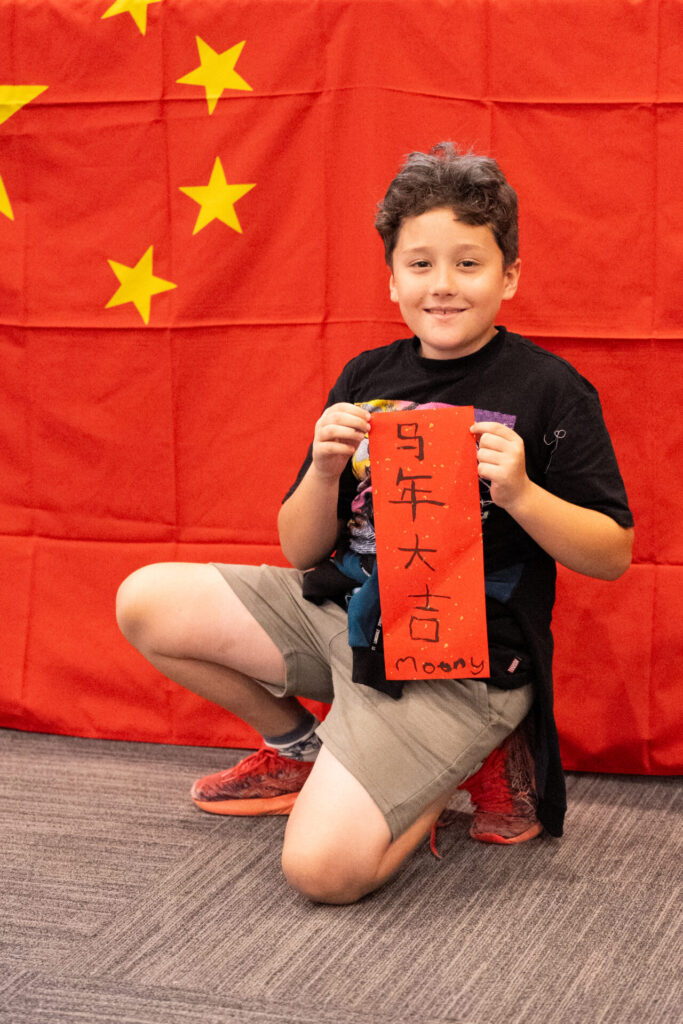 A smiling child kneels in front of a Chinese flag, holding a red paper with Chinese characters and the name Moony written on it. The child wears a black shirt and khaki shorts.