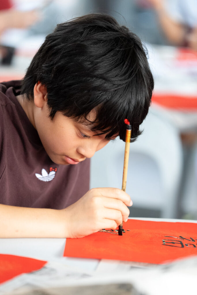 A young boy with black hair concentrates whilst using a calligraphy brush to write black characters on a red sheet of paper at a table.