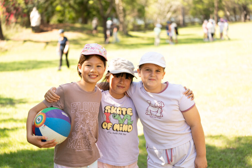 Three children stand arm-in-arm in a sunny park, smiling at the camera. One holds a colourful ball. Other people are visible in the blurred background on the grass.