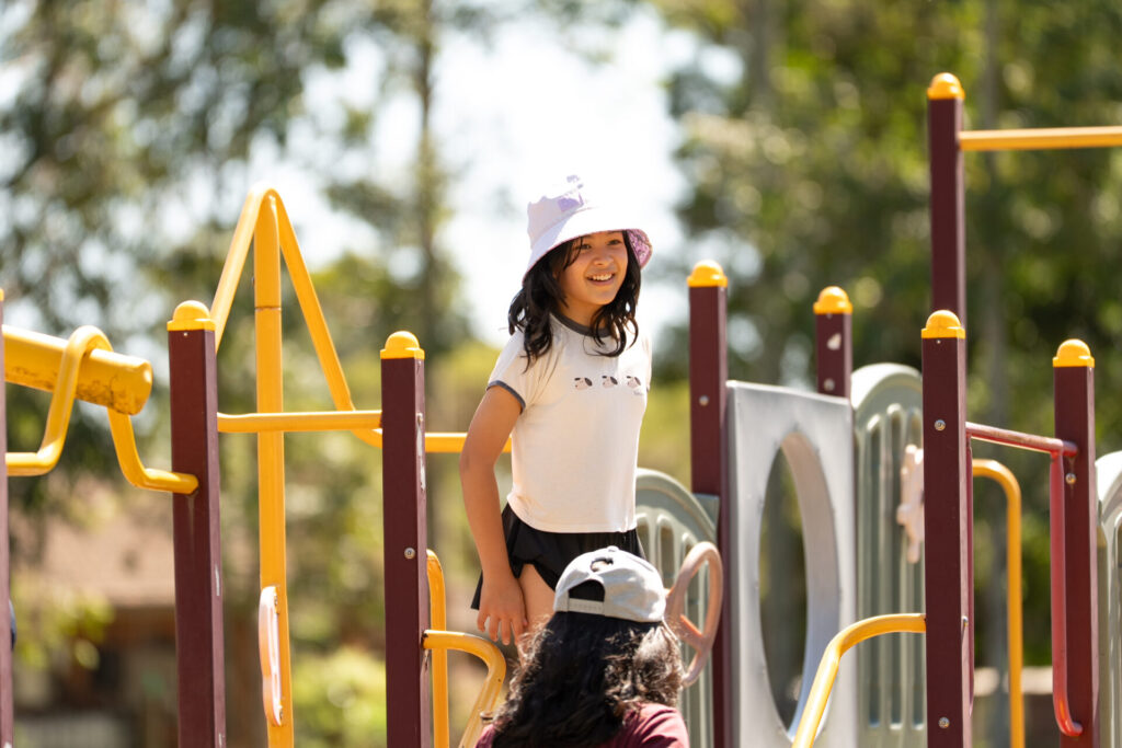 A girl wearing a white hat and T-shirt smiles whilst standing on a colourful playground structure, with another person in a hat nearby. Trees and blurred greenery are visible in the background.
