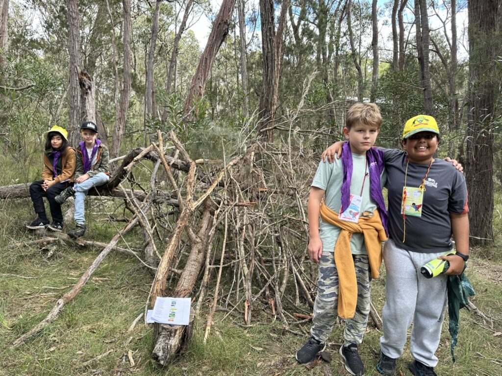 Four children in scouting uniforms stand in a forest next to a shelter made from branches. Two smile and pose in front, while two others sit on logs nearby. Trees and greenery surround them.