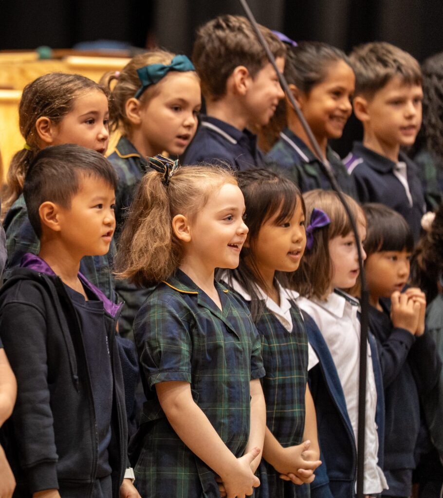 A group of young children in school uniforms stand in rows, joyfully singing together on stage during a music celebration. They appear happy and engaged, with some smiling and looking towards the audience.