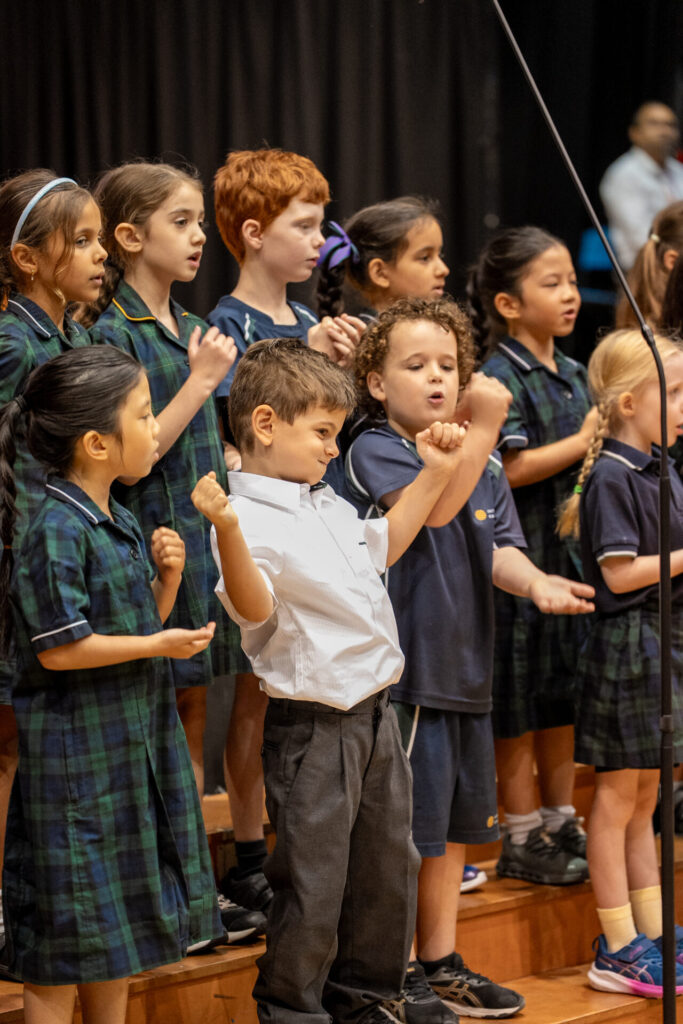 A group of young children in school uniforms stand on staging, singing and gesturing with their hands during a musical performance, celebrating their early years, with a microphone visible in front of them.