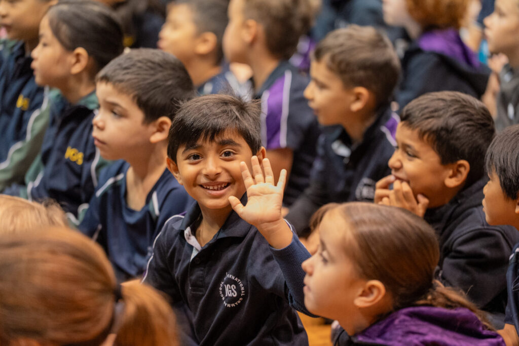 A young boy smiling and waving at the camera whilst sitting among a group of children in school uniforms during an early years joyful celebration. The other children are focused on something out of view.
