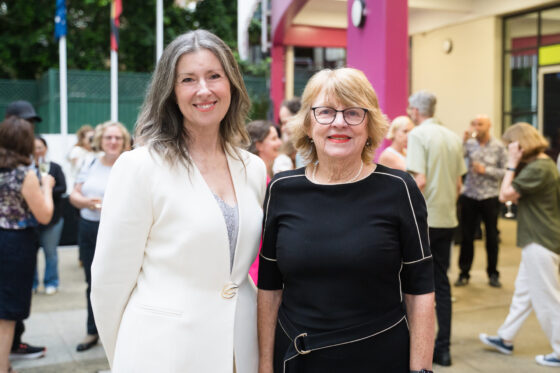 Two women smile for the camera at an outdoor event, radiating talent and joy. One wears a white blazer, the other a black dress. People mingle in the background, with flags waving and a pink pillar adding momentum to the lively scene.