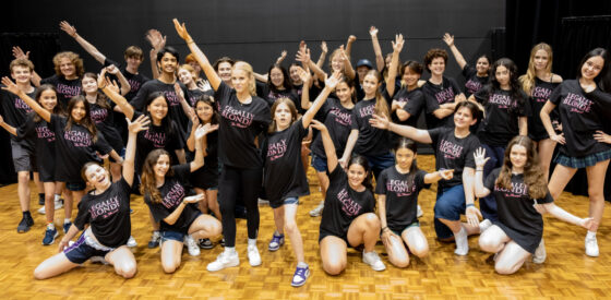 A large group of excited teenagers and a few adults pose together indoors, wearing matching “Legally Blonde” T-shirts. They stand on a wooden floor, smiling and waving during rehearsals, capturing the cheerful, theatrical spirit of Elle Woods at Harvard.