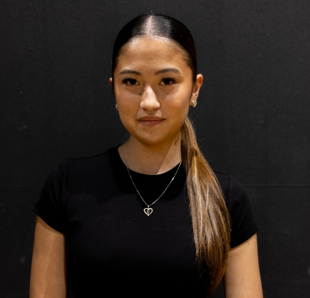 A woman with straight long brown hair in a low ponytail stands against a black background, resembling Elle Woods at Harvard, wearing a black short-sleeved top, heart-shaped necklace, and stud earrings, looking at the camera with a neutral expression.