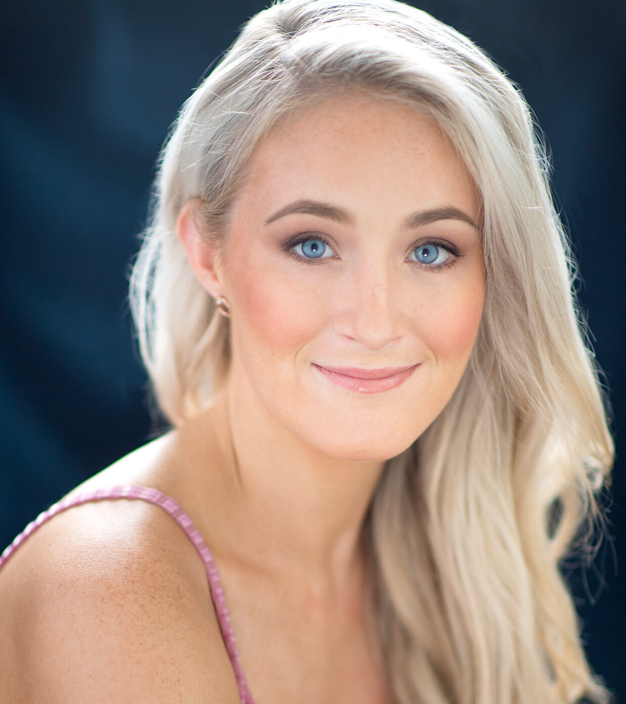 A woman with long blonde hair and blue eyes, styled like Elle Woods, wears a pink sleeveless top and smiles softly at the camera against a dark blurred background, as if ready for rehearsals at Harvard.