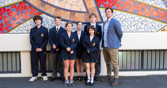 Seven people, six students in school uniforms and one adult in a suit jacket, stand smiling in front of a colourful mosaic wall outdoors. The Mock Trial Team is posed together on a paved area, celebrating their recent victory.