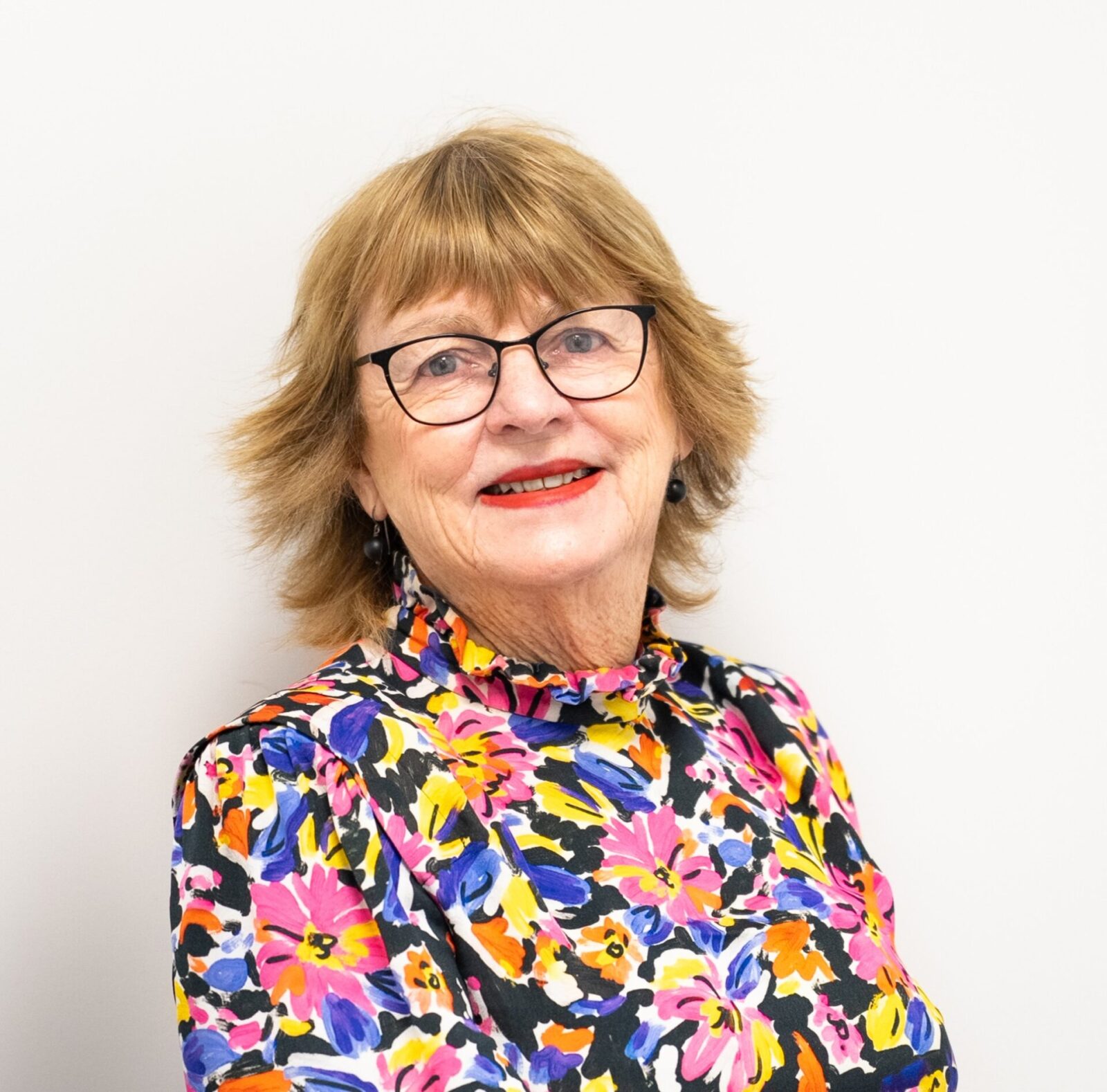 An older woman with short light brown hair, glasses, and red lipstick smiles confidently in a colourful floral blouse, posing for her School Board portrait against a plain white background.