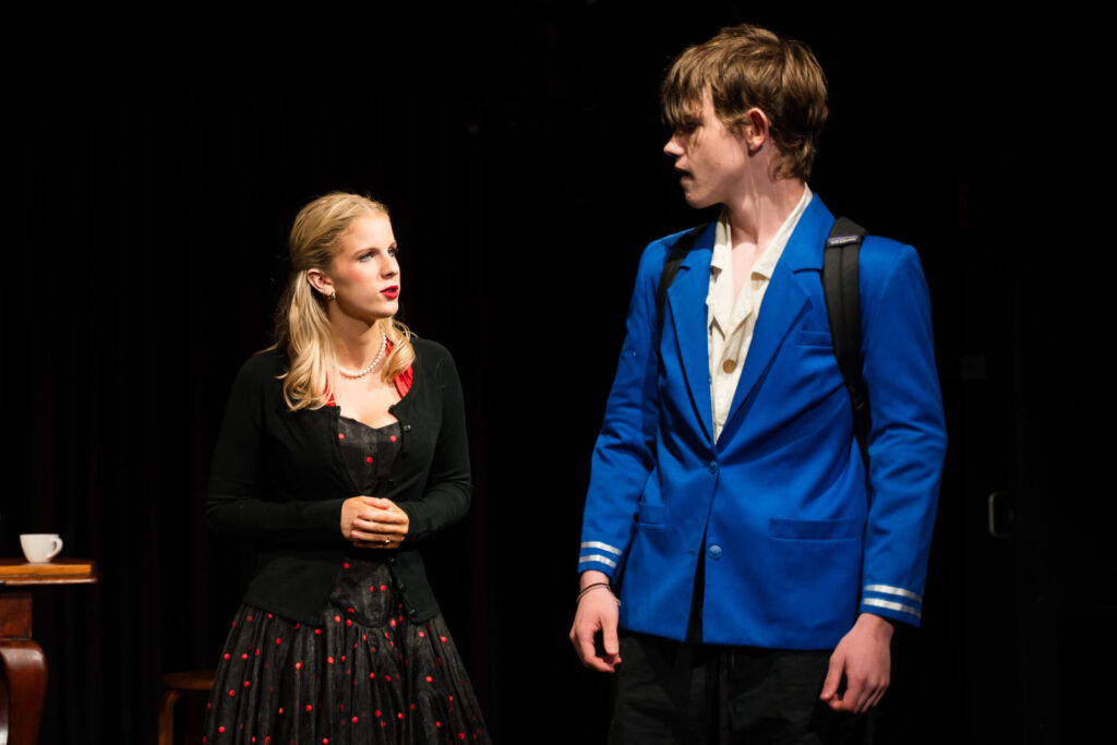 A young woman in a black dress with red polka dots and a young man in a bright blue jacket stand on stage, looking at each other under dramatic lighting, with a table and teacup visible in the background.