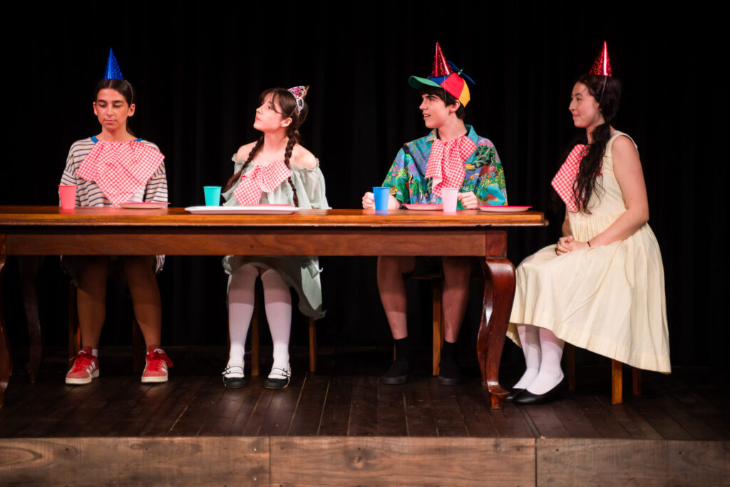 Four people wearing party hats sit at a wooden table with serviettes tucked into their collars, looking to the left. Colourful cups and plates are on the table, and the background is a dark stage curtain.