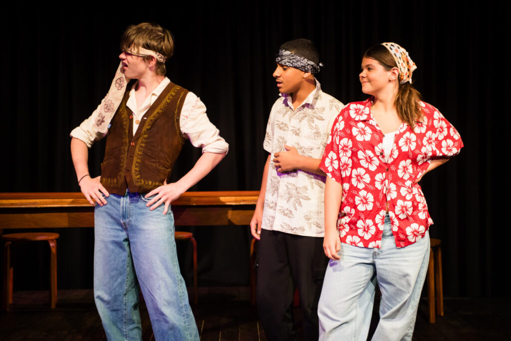 Three people stand on stage in bright costumes, including floral shirts and headscarves, with a wooden table and chairs behind them. They appear to be performing in a play, looking offstage with expressive poses.