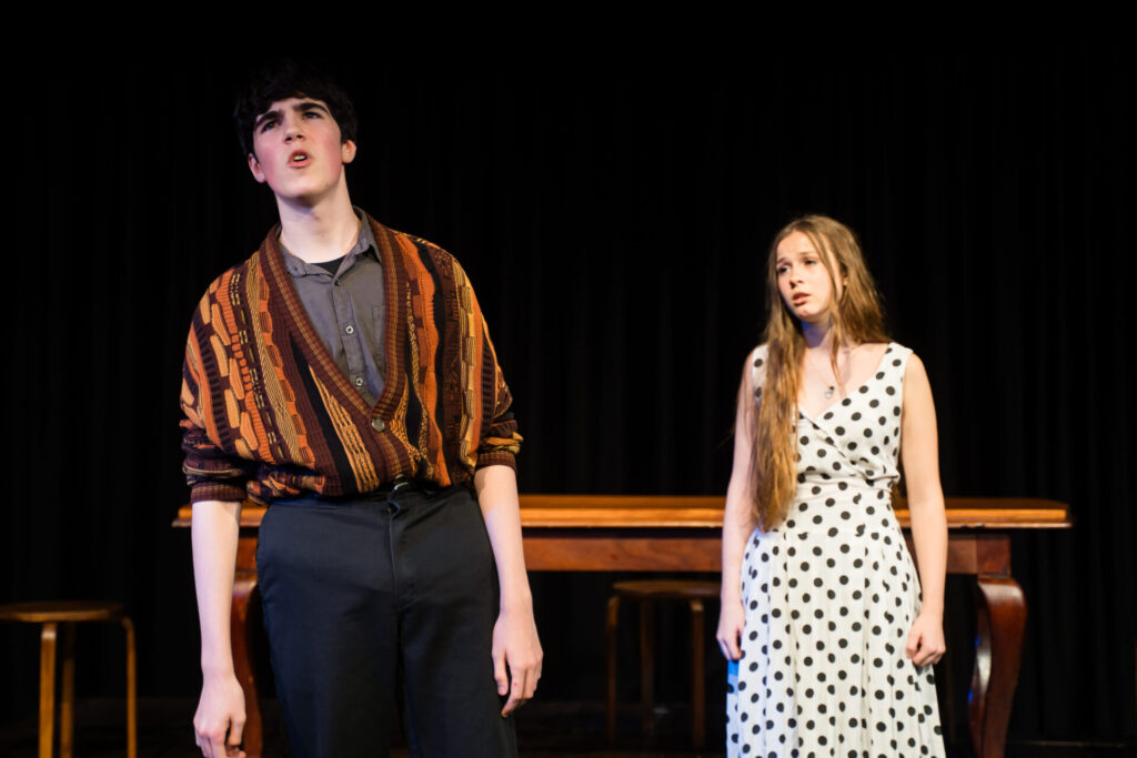 A young man in a patterned jumper stands in the foreground, speaking passionately, while a young woman in a white spotted dress stands behind him, looking sad. They are on a stage with a table and chairs in the background.