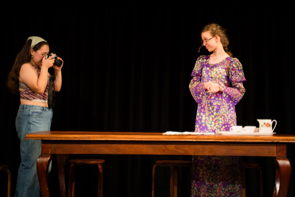 Two young women are on stage. One is taking a photo with a camera, wearing a headband and jeans. The other, in a long floral dress, stands by a wooden table set with cups and a teapot, with a black backdrop behind them.