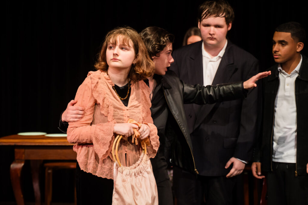 Four young people in formal clothing stand together on stage. One person in a pink ruffled blouse holds a bag and looks away whilst another in black comforts them, arm round their shoulders. Two others stand close by, watching.