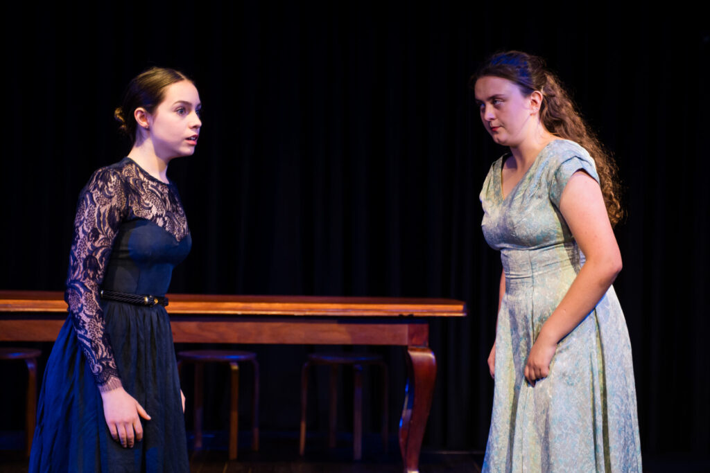 Two young women in dresses stand facing each other on stage, appearing tense and emotional. A wooden table and two stools are in the background, and the lighting emphasises their expressions against a dark backdrop.