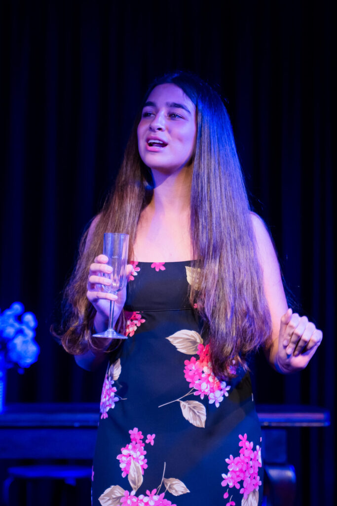 A young woman with long brown hair, wearing a sleeveless black dress with pink and white floral patterns, holds a champagne glass and gestures whilst speaking on a stage with dark curtains in the background.