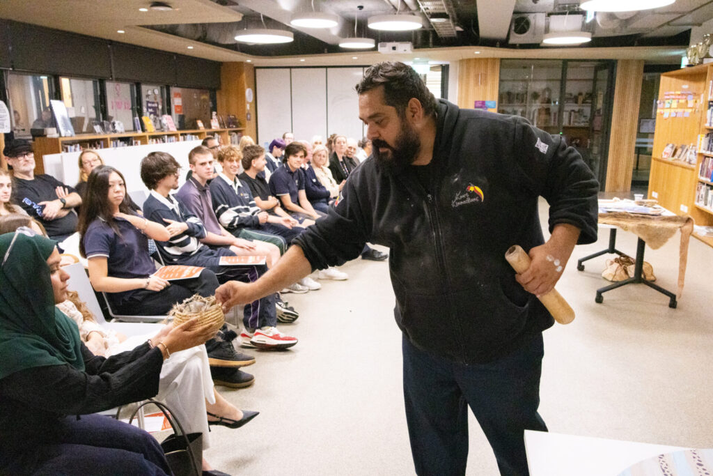 A man holding a wooden stick hands a basket to a seated woman in a classroom, as pupils watch attentively—an exchange echoing the spirit of "Always Was, Always Will Be.