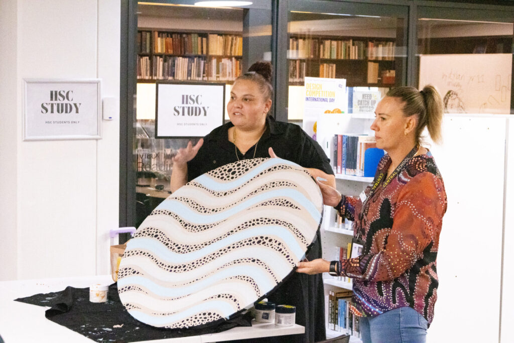 Two women stand in a library, one speaking while the other holds a large, round artwork with wavy patterns and dot designs. Shelves of books and HSC STUDY signs are visible, celebrating the message: Always Was, Always Will Be.