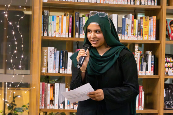 A woman in a green hijab and black outfit holds a microphone and papers, smiling as she speaks in front of bookshelves in a library decorated with fairy lights, beneath a banner reading 