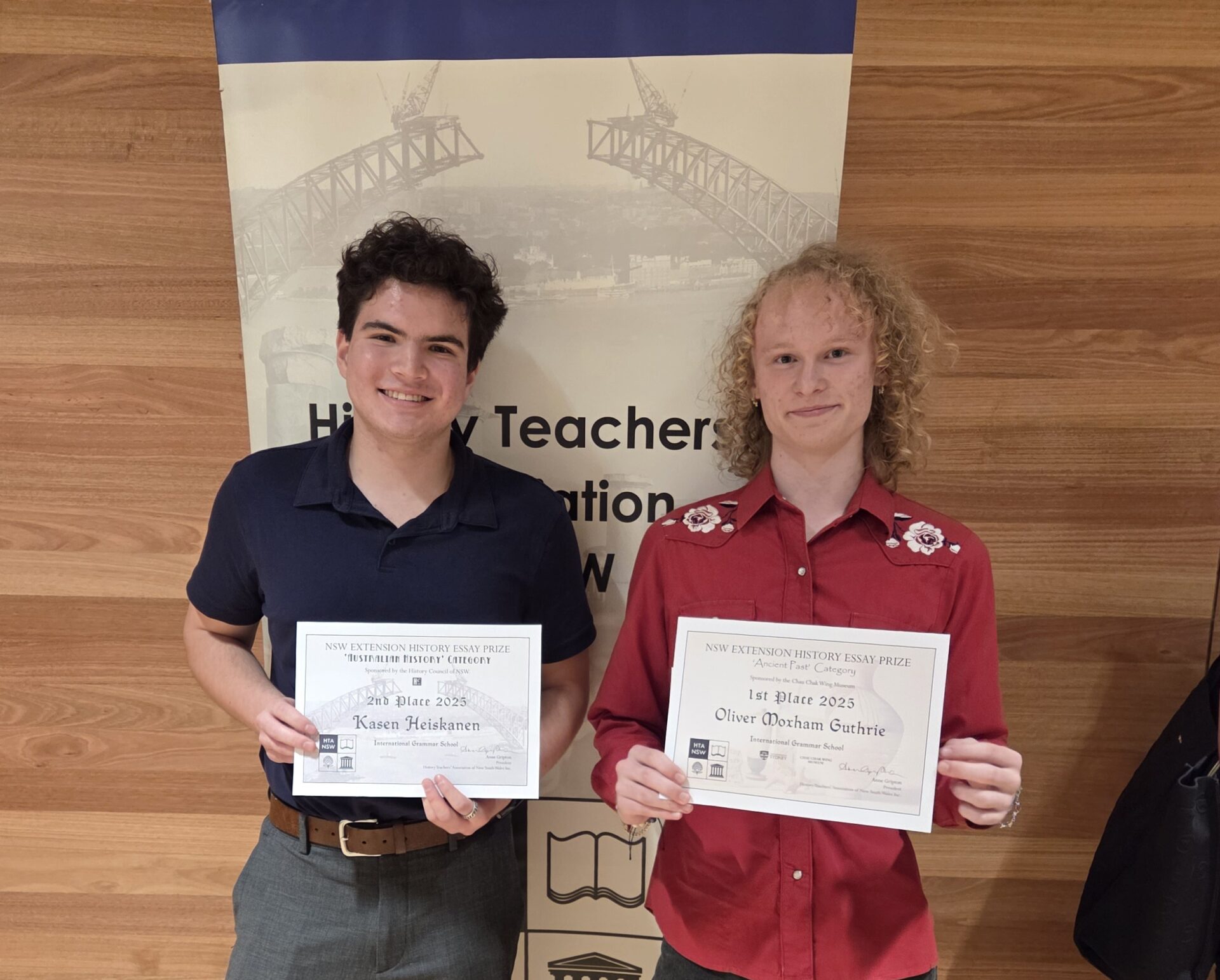 Two young high achievers stand side by side, smiling and holding certificates for winning first place in a 2023 history essay competition, against a wooden wall and a banner displaying a bridge illustration.