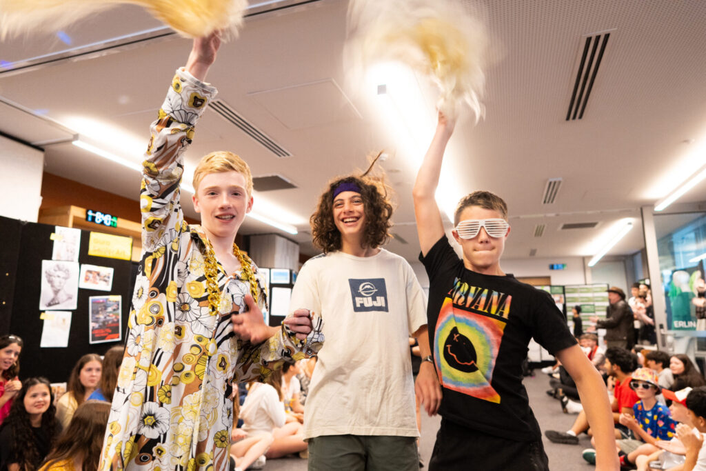 Three boys stand indoors at a lively ArtsFest event. One waves pom-poms in a floral pyjama top, another sports curly hair and a headband, and the third rocks a Nirvana shirt and shutter shades—a true journey through time at IGS.