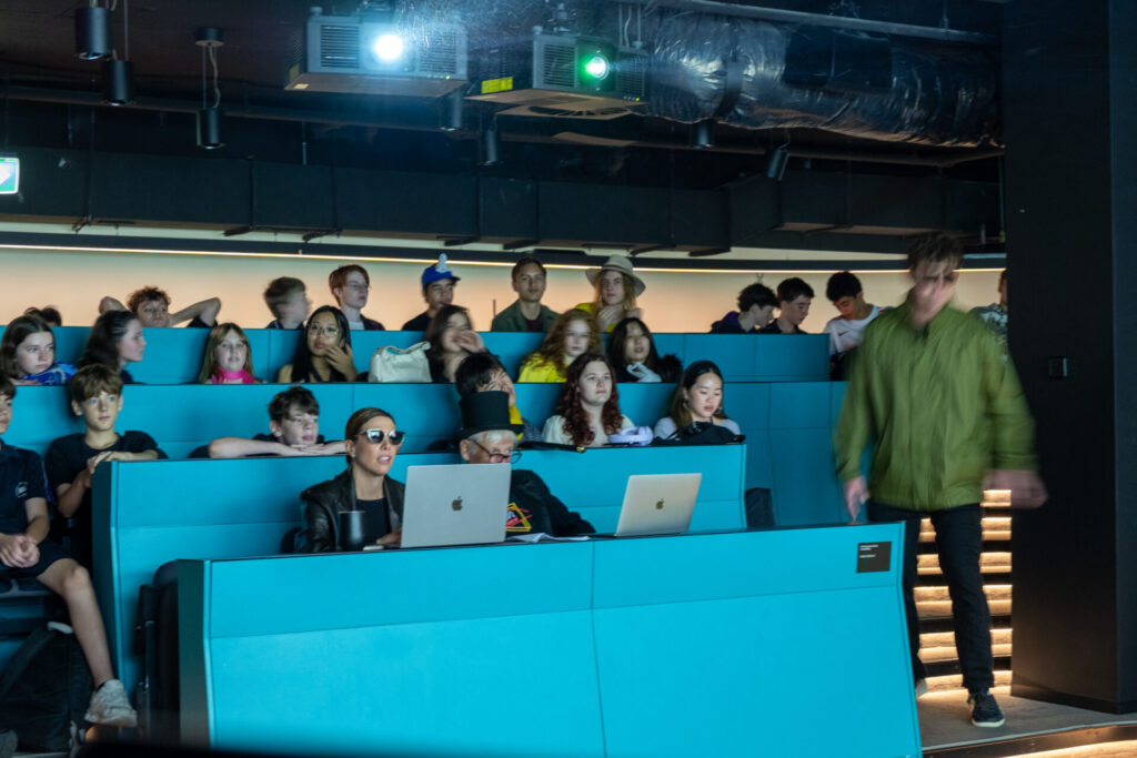 A group of students sits in tiered, bright blue lecture theatre seats, some with laptops open, during IGS. One person in the front row wears sunglasses while a student in a green jacket walks past, blurred—a snapshot from their journey through time.
