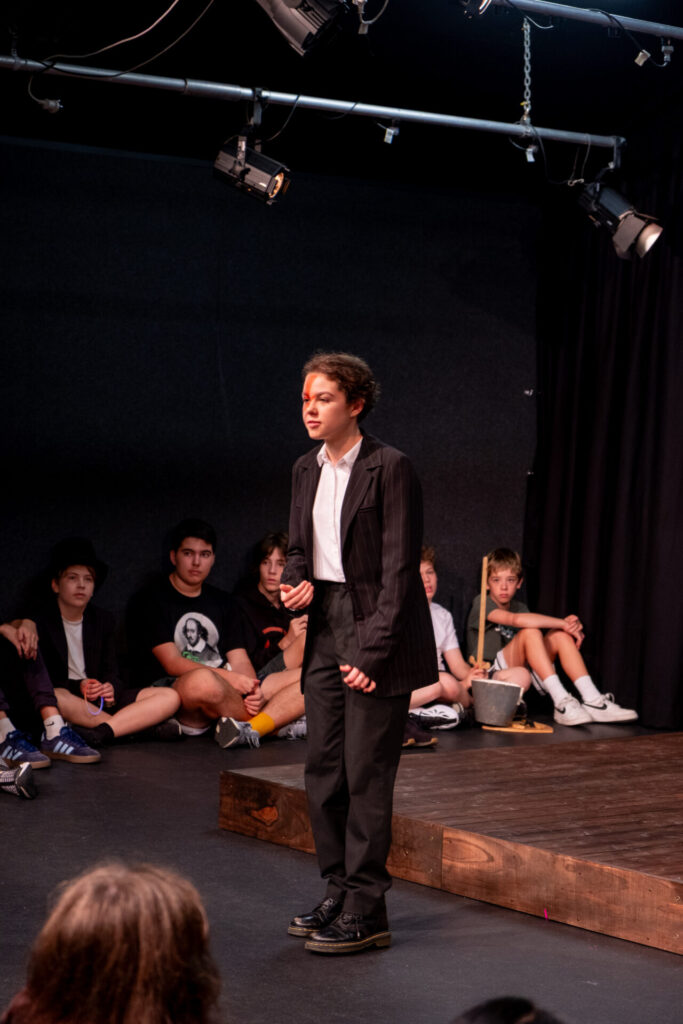 A young person in a black suit and white shirt stands and speaks on stage during IGS ArtsFest, while children watch from the floor. Stage lights shine above, evoking a journey through time in this lively theatre performance or rehearsal.