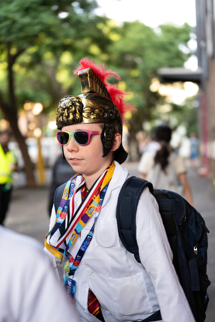A boy wearing a Roman-style helmet with a red plume, pink sunglasses, a white lab coat, and colourful lanyards stands outdoors at ArtsFest IGS, ready for a journey through time. Trees and other people are visible in the background.