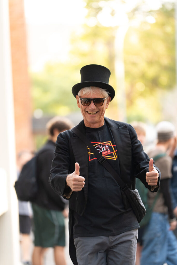 An older man wearing a black top hat, sunglasses, and a black jacket gives two thumbs up whilst smiling outdoors at ArtsFest, capturing the spirit of a joyful journey through time. A blurred crowd and trees are visible in the background.