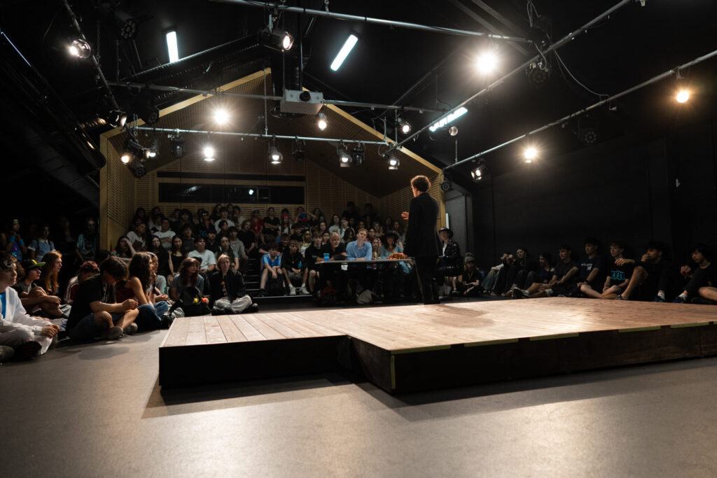 A person stands on a stage at IGS ArtsFest in a dark theatre, facing an audience seated closely together and attentively watching. Bright stage lights illuminate their journey through time.
