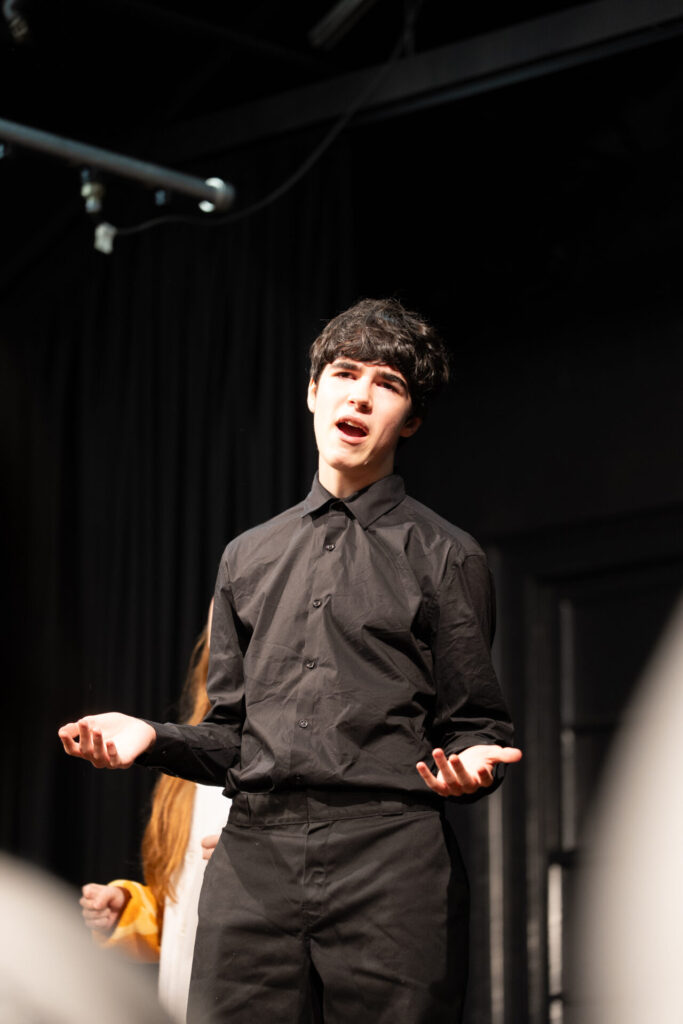 A young person with short dark hair, dressed in a black shirt and trousers, stands on stage with arms outstretched, passionately performing at ArtsFest, framed by a dark curtain backdrop—a moment in their journey through time.