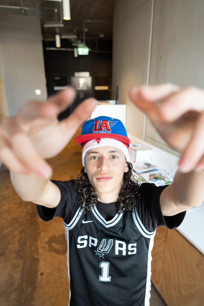 A person wearing a San Antonio Spurs jersey and a blue-and-red LA cap stands indoors at IGS ArtsFest, reaching both hands towards the camera with a focused expression, as if inviting you on a journey through time.