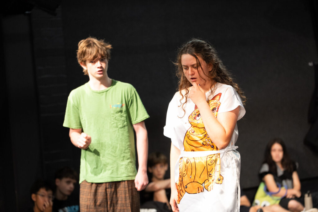 Two teenagers perform on stage at ArtsFest; the boy in a green shirt and brown shorts raises his fist whilst the girl in a white costume with an orange design, part of IGS’s journey through time, looks distressed, touching her neck as others watch in the background.