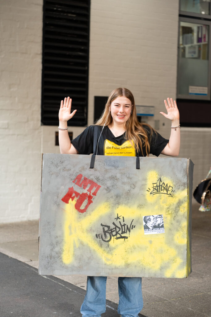 A young woman stands outside at ArtsFest with her hands raised, wearing a large cardboard costume painted to look like a graffiti-covered wall. She is smiling and dressed casually in jeans and a black T-shirt, embodying a creative journey through time.