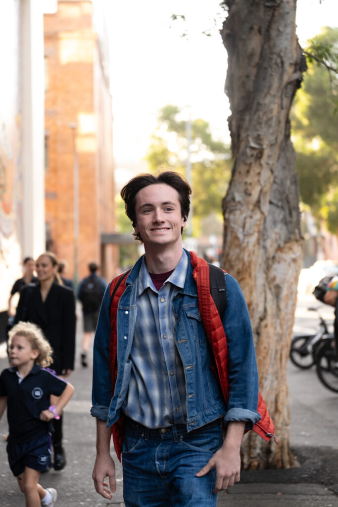 A young man with dark hair smiles while walking on a city pavement during ArtsFest. He wears a denim jacket, checked shirt, red jacket over his shoulders, and a rucksack. People, including a child, walk nearby amidst trees and buildings.