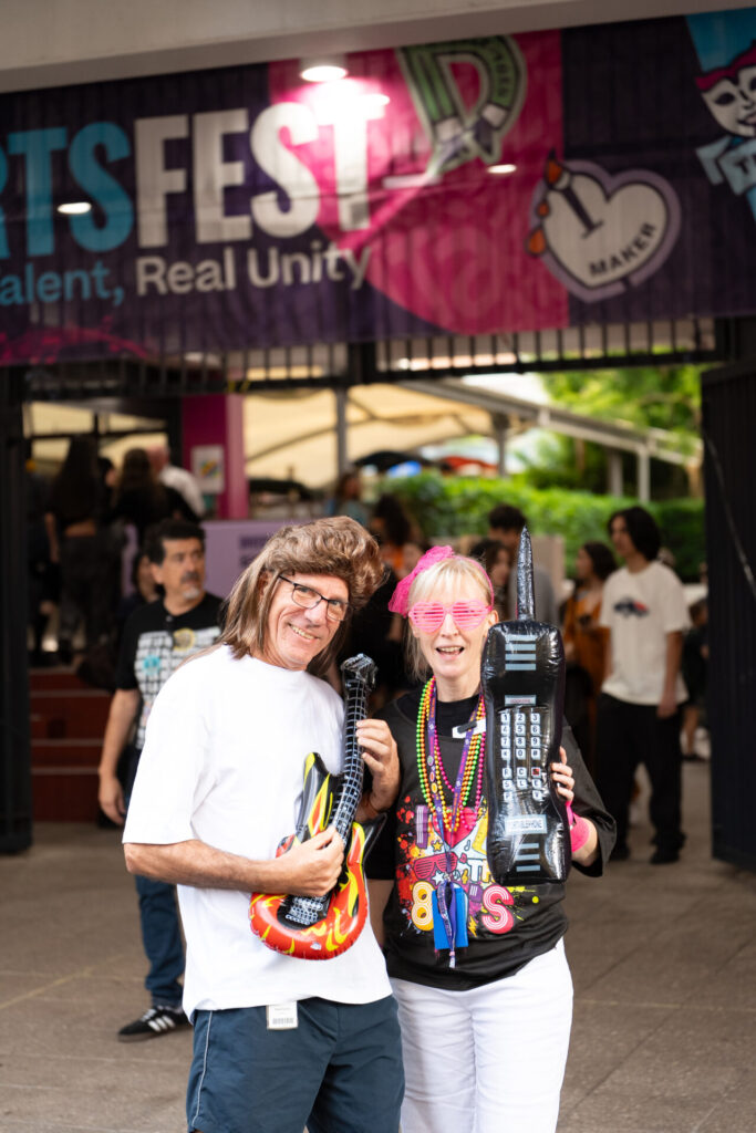 Two people in colourful 1980s-themed outfits and wigs pose with inflatable guitars, smiling in front of an ArtsFest banner at an outdoor event—a fun journey through time at IGS, with other attendees enjoying the festivities in the background.
