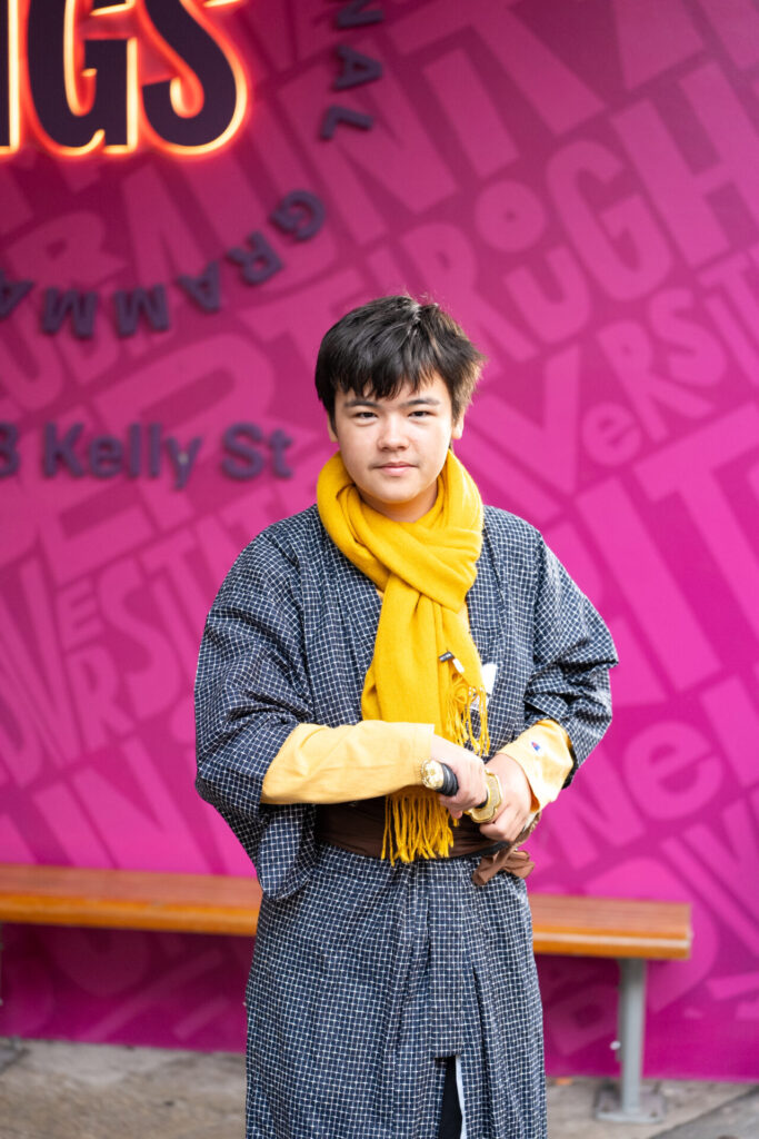 A person wearing a yellow scarf and dark patterned dressing gown stands in front of a vibrant pink wall with bold lettering, capturing the spirit of ArtsFest. They hold an object in their hand, looking directly at the camera; a wooden bench rests behind them.