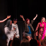 Five people stand on stage at IGS ArtsFest, smiling and cheering with arms raised, facing an applauding audience. Their colourful outfits—including a shiny pink suit and a hijab—capture the joyful, celebratory spirit of this journey through time.
