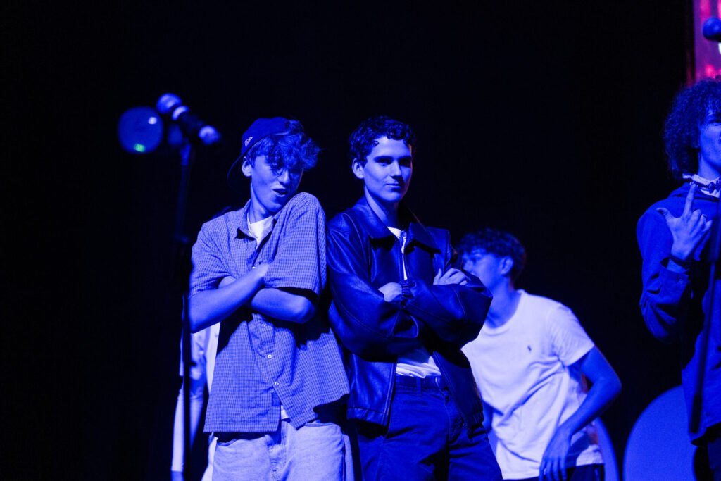 Two young men stand on stage at ArtsFest, arms crossed under blue lighting—one in a cap and checked shirt, the other in a leather jacket—as they embark on a journey through time. Others from IGS are visible in the background with microphones nearby.