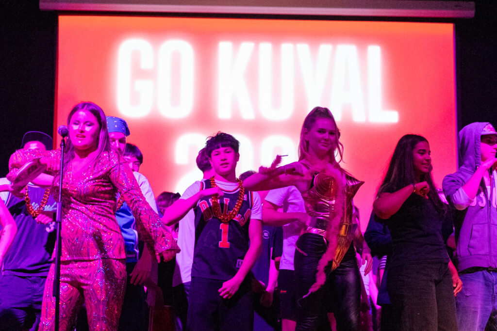 A group of people stand on stage under pink lighting, some wearing costumes, in front of a large screen displaying the words GO KUVAL in bold white letters during the IGS ArtsFest—a vibrant journey through time.