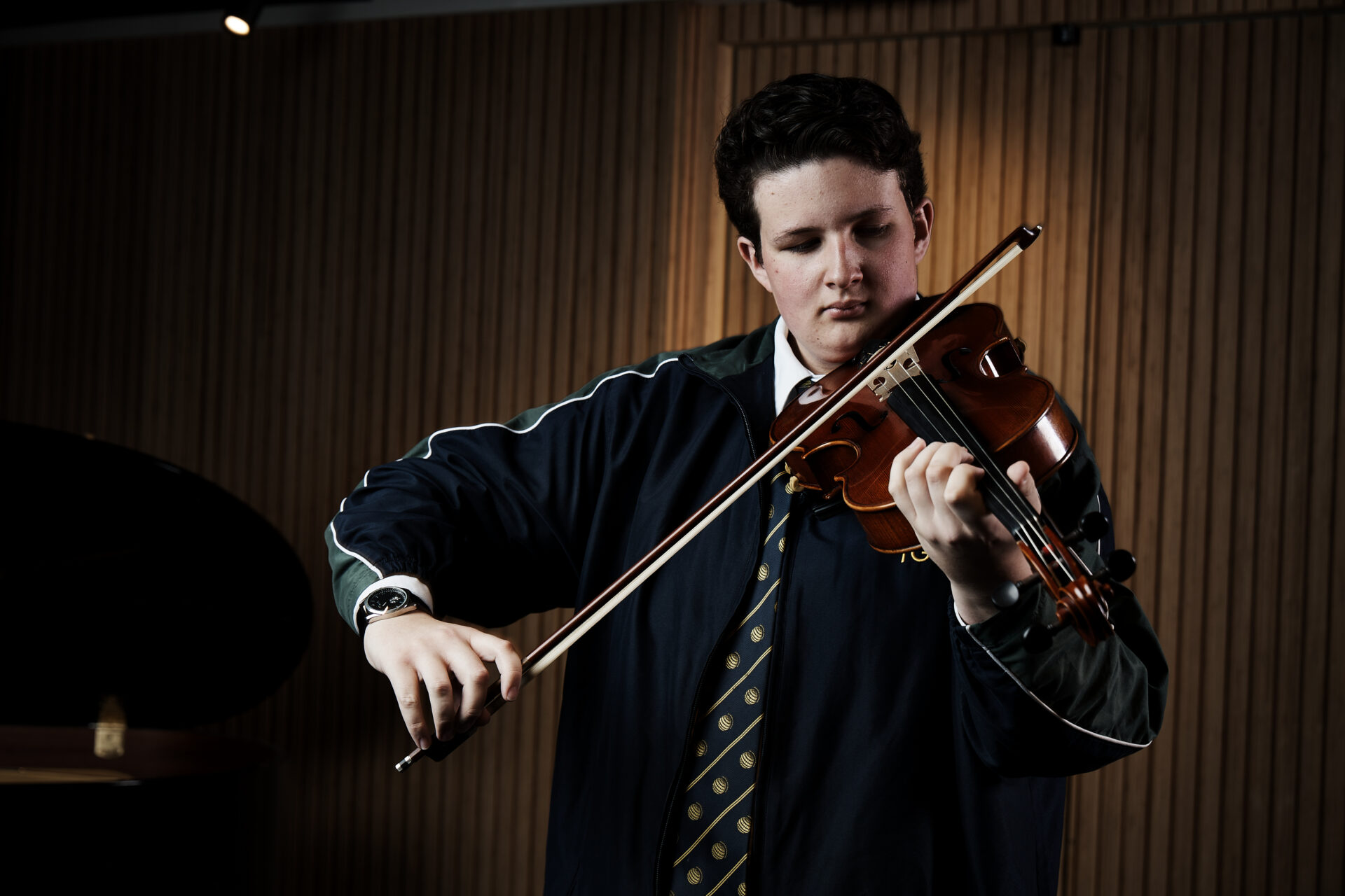 A young person wearing a blue jacket and patterned tie plays a violin with focus, standing indoors against vertical wooden panels—an inspiring moment supported by the IGS Foundation.